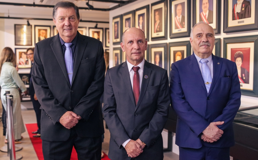 Três homens de terno, em pé, sorriem. Eles estão à frente de uma galeria de quadros