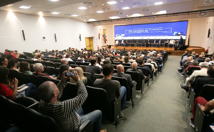 Um evento ocorre em um auditório. No palco, a frente, estão sentadas dez pessoas enquanto uma fala, em pé, em um púlpito. ao fundo, há um telão. Há várias pessoas sentadas em cadeiras pretas enfileiradas voltadas para o palco.