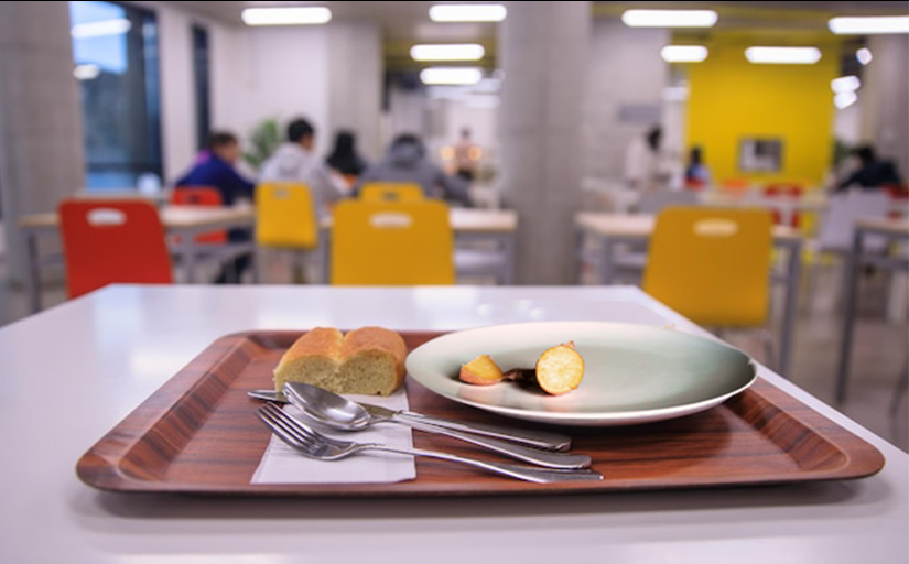 Bandeja sobre a qual há um prato com pouca comida e dois pães pequenos ao lado. Ao fundo, desfocado, um refeitório