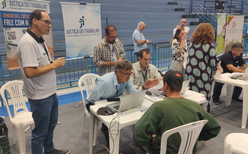 Foto de um atendimento feito por um homem a outro homem, em mesas de plásticos, dentro de um ginásio