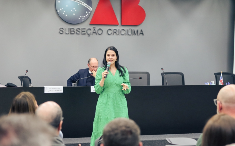 Uma mulher de vestido verde fala ao microfone para uma plateia 