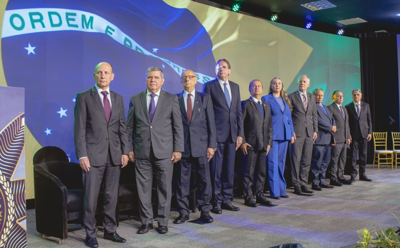 Grupo de autoridades em pé, lado a lado, em frente à imagem projetada da bandeira do Brasil, em evento formal.
