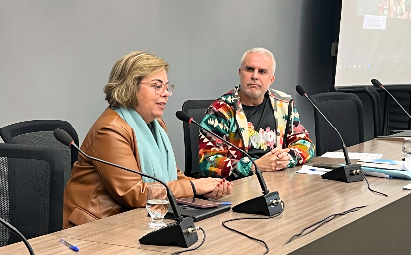 Uma mulher e um homem estão sentados em um palco fazendo uma palestra