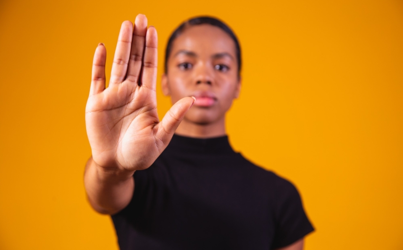 Uma mulher negra está com a mão estendida para frente, com rosto sério e vestida de preto