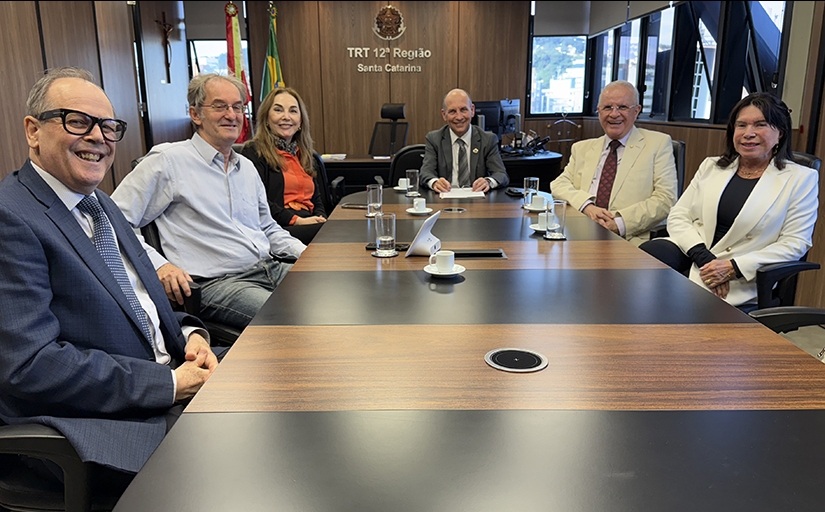 Quatro homens e duas mulheres sentados em torno de uma mesa retangular sorriem para a foto, em uma sala com janelas abertas