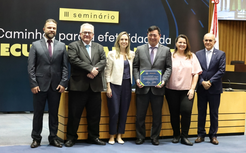 Quatro homens de terno, em pé, e duas mulheres vestidas formalmente posam para foto em um auditório. Um dos homens mostra um certificado.