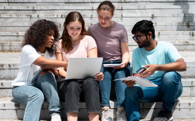 Grupo de quatro estudantes sentados em escadaria ao ar livre, olhando para um notebook com atenção.
