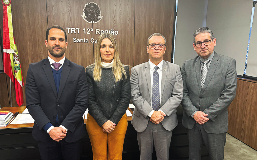 Quatro pessoas posam lado a lado em frente ao letreiro “TRT 12ª Região – Santa Catarina”, dentro de um gabinete. À esquerda, bandeira de Santa Catarina. Todos estão com trajes formais.