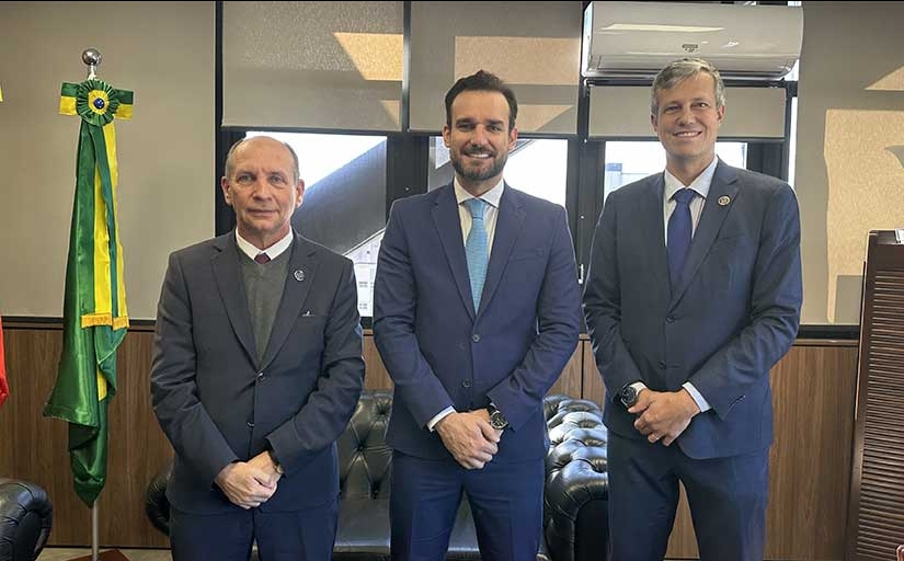 Três homens de terno, lado a lado, posam para foto, dentro de uma sala. À esquerda, aparece a bandeira do Brasil