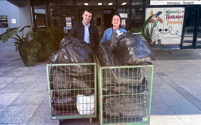 Imagem de dois carrinhos de transporte recheados de sacos pretos com artigos dentro. Um homem e uma mulher sorriem para a foto 