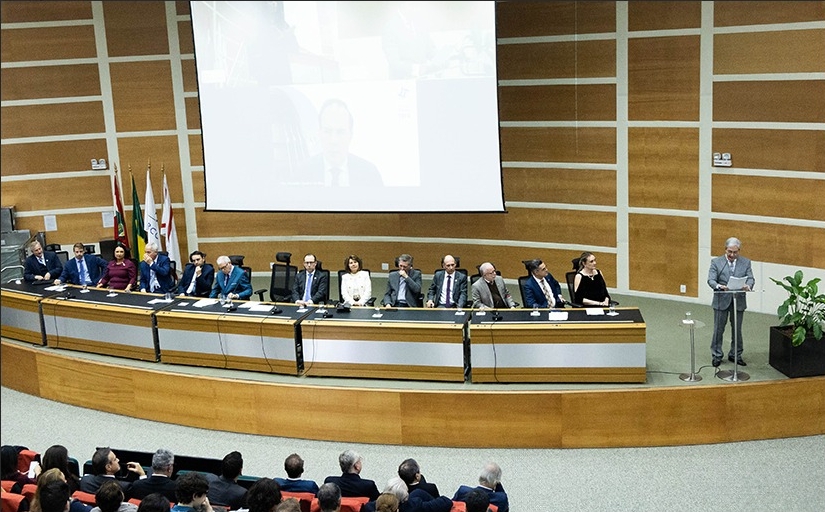 Cerimônia formal em auditório. Autoridades sentadas à mesa no palco, homem discursa no púlpito. Público assiste. Telão ao fundo exibe participante remoto. Bandeiras à esquerda, planta decorativa à direita.