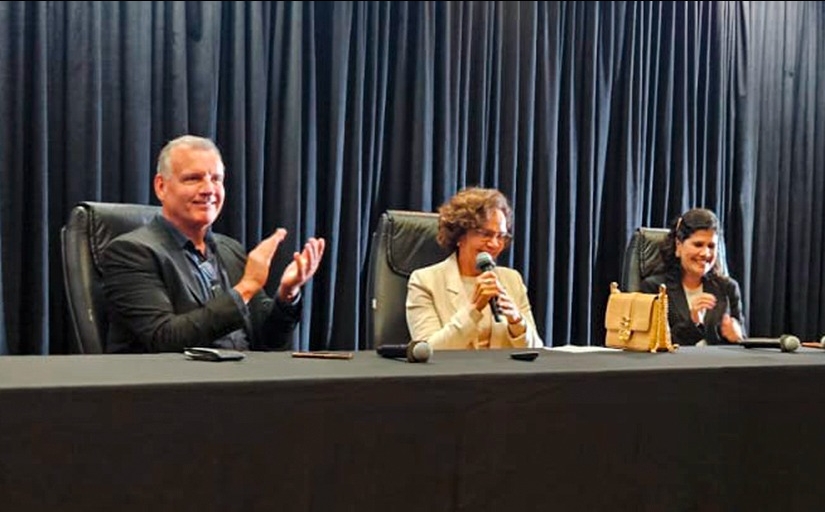 Um homem e duas mulheres estão sentados em um palco dando uma palestra