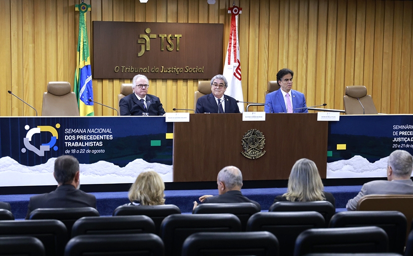 Três homens de terno sentados em uma mesa em um evento.