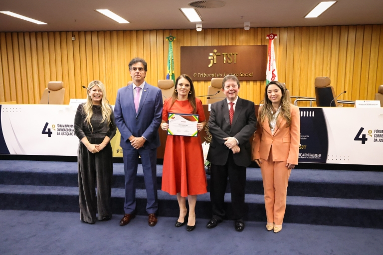 Fotografia na qual cinco pessoas posam em auditório do TST; ao centro, mulher segura certificado
