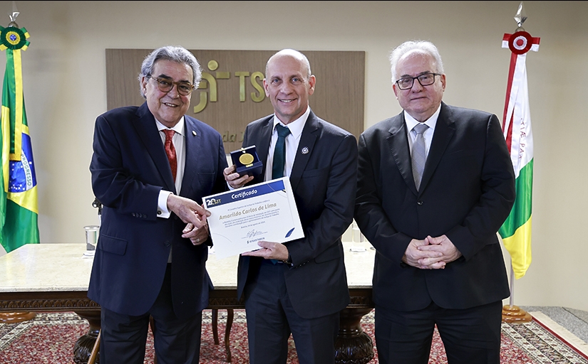 Três homens de terno e gravata estão em pé, posando para a foto. O homem que estão no centro segura um certificado e uma medalha