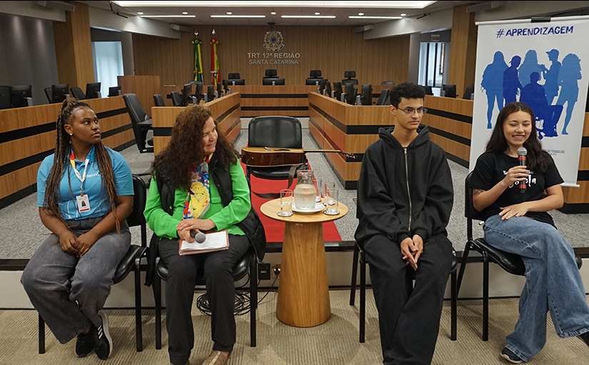 Três jovens e uma mulher madura sentados em cadeiras, dentro de uma ampla sala de julgamentos. Uma das jovens segura o microfone e sorri