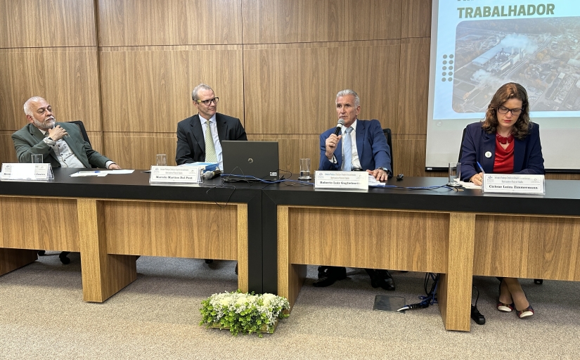 Três homens e uma mulher sentados à mesa de um auditório. Um dos homens fala ao microfone