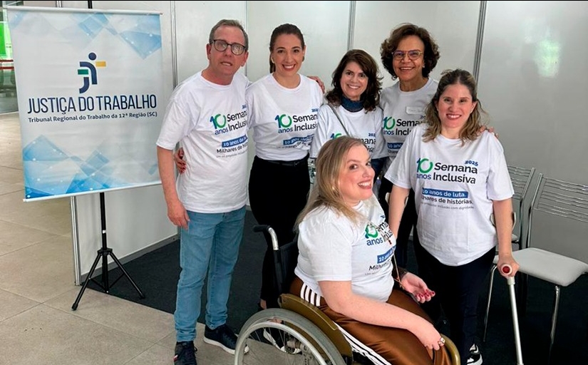 Um homem e quatro mulheres em pé e uma mulher na cadeira de rodas estão posando para a foto. Ao lado deles um banner escrito: Justiça do Trabalho