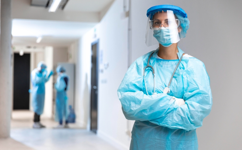 Foto de uma profissional de saúde vestida com equipamentos de proteção individual, em um ambiente hospitalar