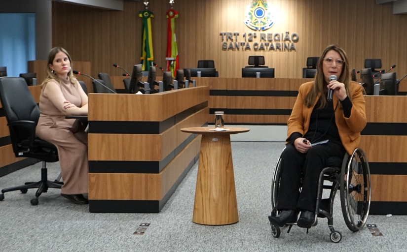 Foto de uma mulher de cadeiras de rodas dando uma palestra. No fundo um painel escrito Tribunal Regional do Trabalho da 12ª Região.
