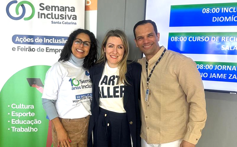 Três pessoas sorriem para foto durante a Semana Inclusiva Santa Catarina, em frente a banner do evento.