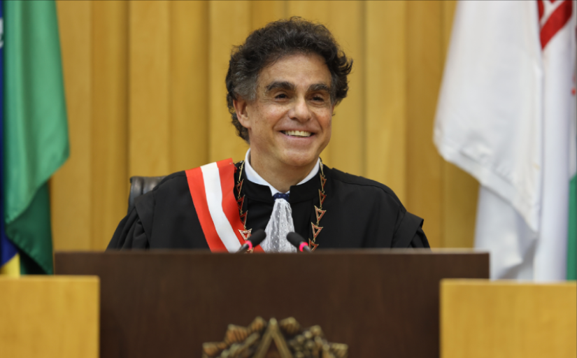 Homem de cerca de 60 anos, vestido com toga de juiz e faixa, sorri 