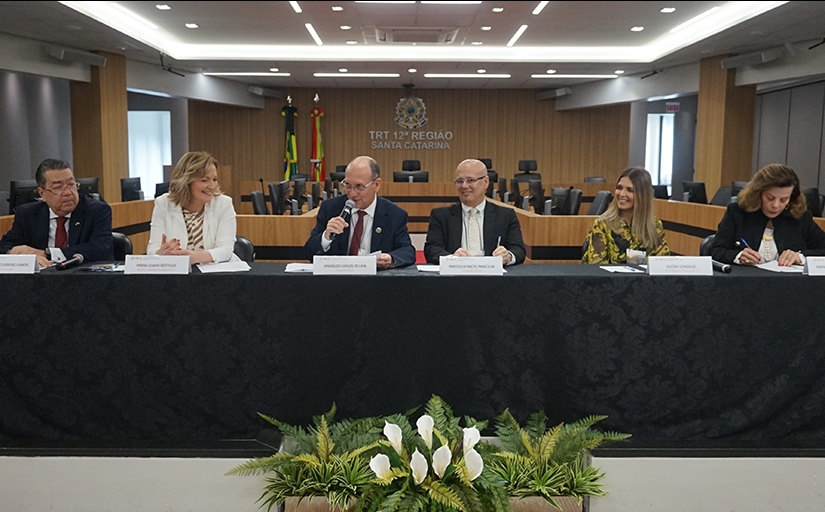 Três homens e três mulheres sentadas à mesa de um evento, coberta por uma toalha preta e um buquê de flores no chão.