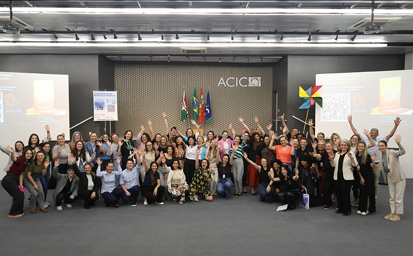 Grupo de pessoas reunidas e sorrindo em auditório da ACIC, com bandeiras ao fundo e clima de celebração.