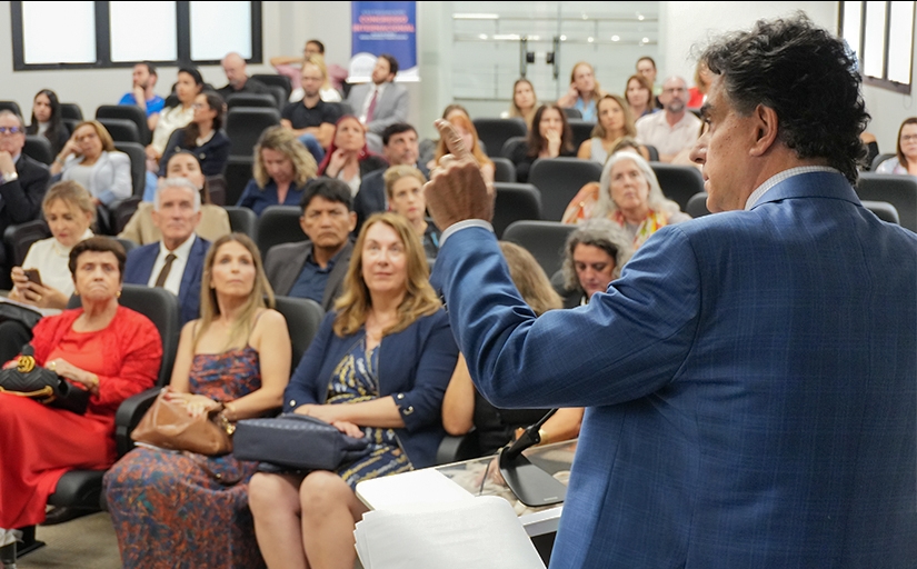 Foto de um homem de costas, de terno, dando uma palestra. Ao fundo o público assistindo