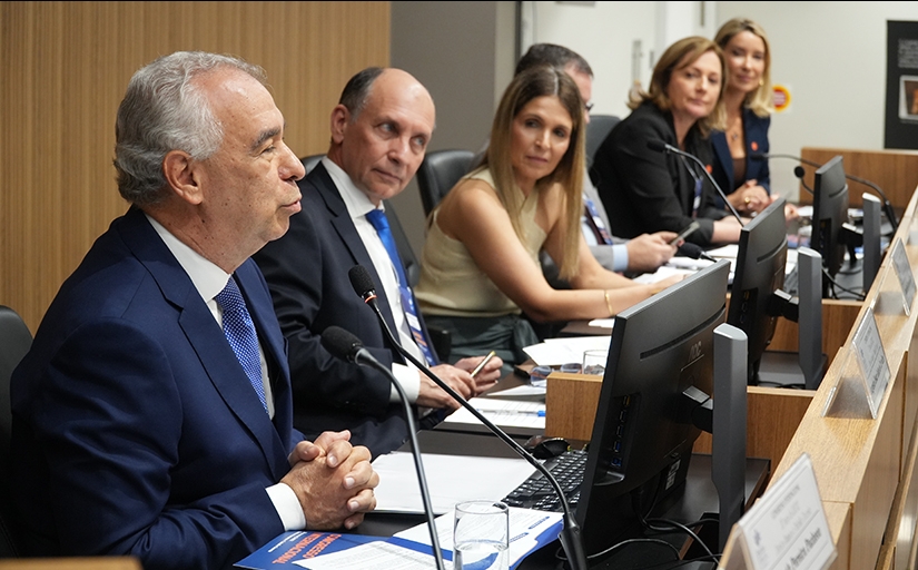 Foto de uma mesa de honra de um evento, com um homem falando e mais homens e mulheres sentados olhando o palestrante