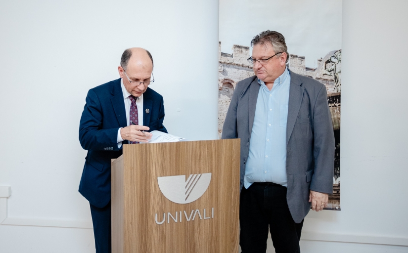 Dois homens estão posicionados ao lado de um púlpito de madeira com o nome e logotipo da UNIVALI. Um deles escreve ou assina um documento sobre o púlpito.