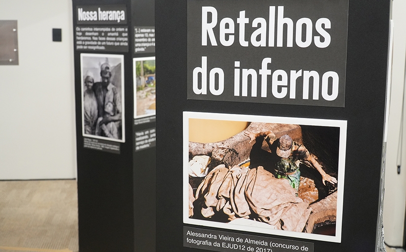 Totem de uma exposição mostrando a foto de um trabalhador no meio de muitos tecidos. O texto acima da foto é "Retalhos do Inferno".