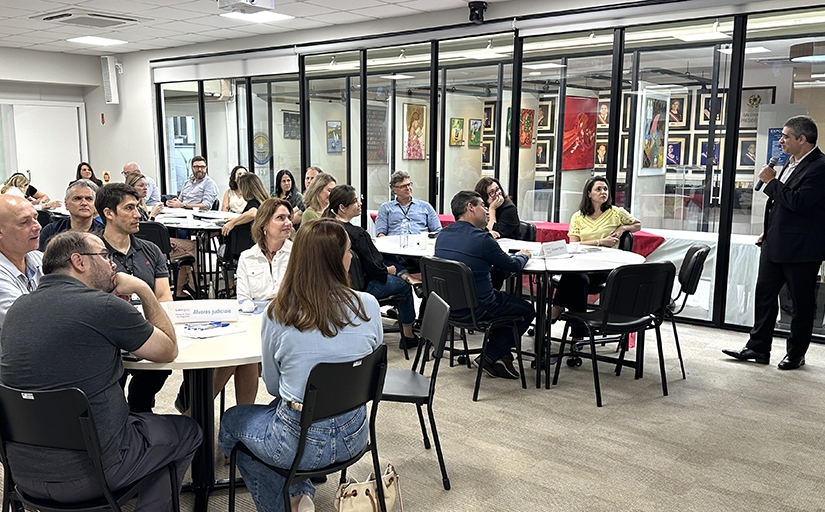 Grupo de pessoas participa de palestra em ambiente corporativo, com mesas redondas e obras de arte expostas ao fundo.