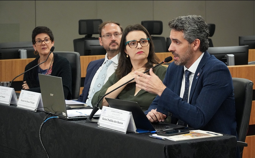 Palestrantes participam de mesa-redonda em auditório, com notebooks e microfones sobre a mesa.