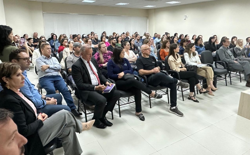 Foto em diagonal mostra cerca de 90 pessoas sentadas em cadeiras dentro de um auditório
