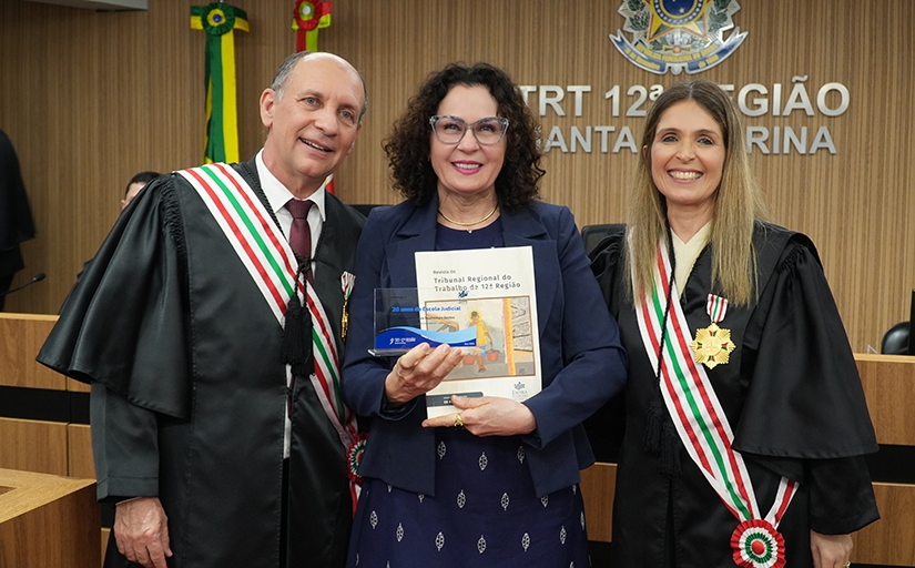 Três pessoas sorridentes posam em solenidade no TRT-12; ao centro, mulher segura troféu e livro comemorativo.