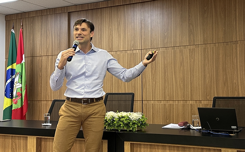 Ao centro, um homem jovem fala ao microfone. Ao fundo dele, um painel de madeira