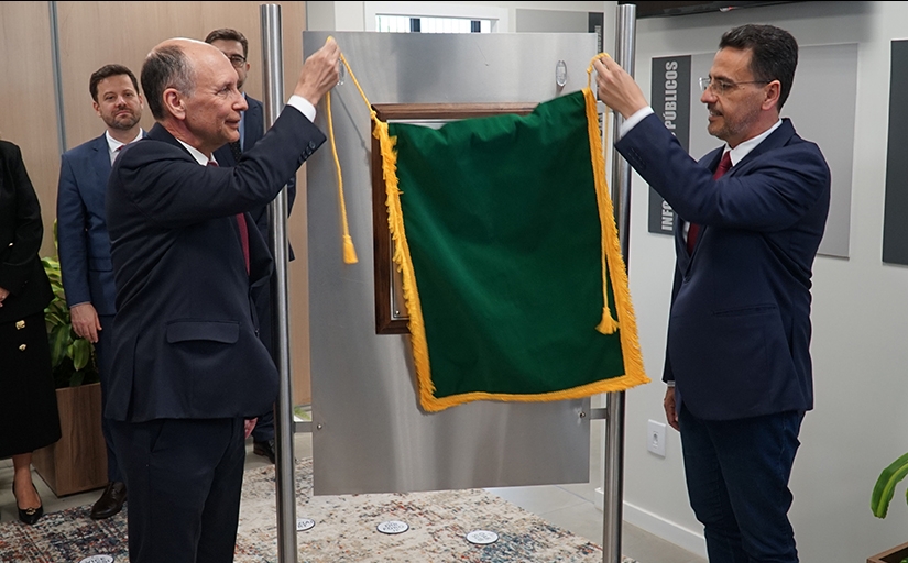 Um homem calvo, de cerca de 60 anos, de terno, e outro homem moreno, também de terno, retirar um pano verde que está cobrindo uma placa de metal