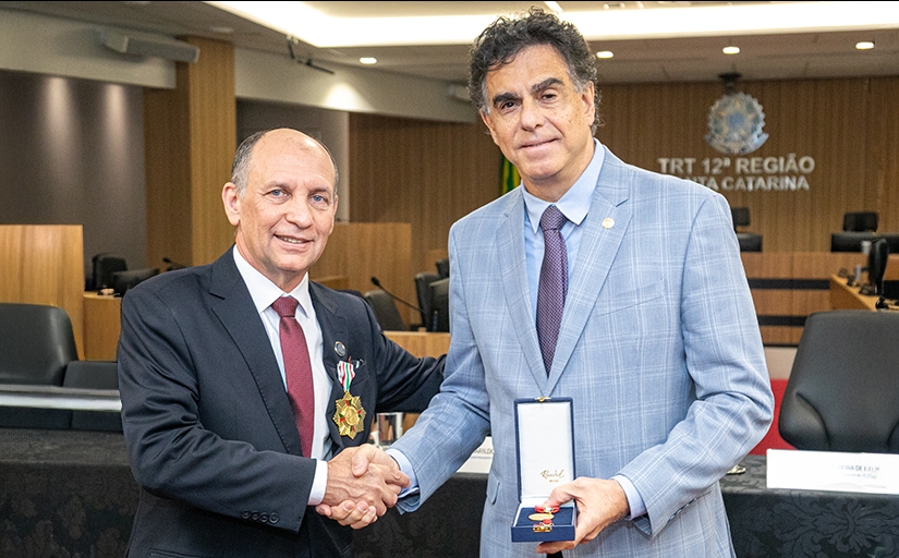 Dois homens de terno, com cerca de 60 anos, se cuprimentam. Um deles, de terno claro, exibe uma caixa contendo uma medalha.