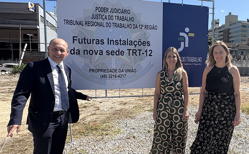 Um homem de terno e duas mulheres ao lado de uma placa de cerca de 2 metros de altura sobre um terreno vazio