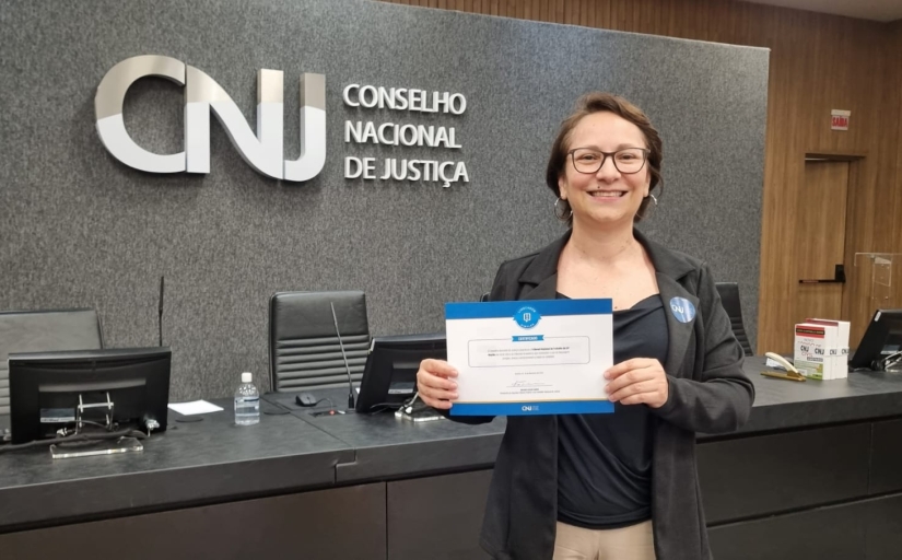 Mulher sorridente segurando o certificado. Ao fundo a logo do CNJ