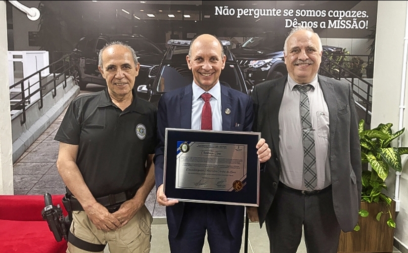 Três homens posam para a foto; ao centro, um deles segura uma placa de homenagem
