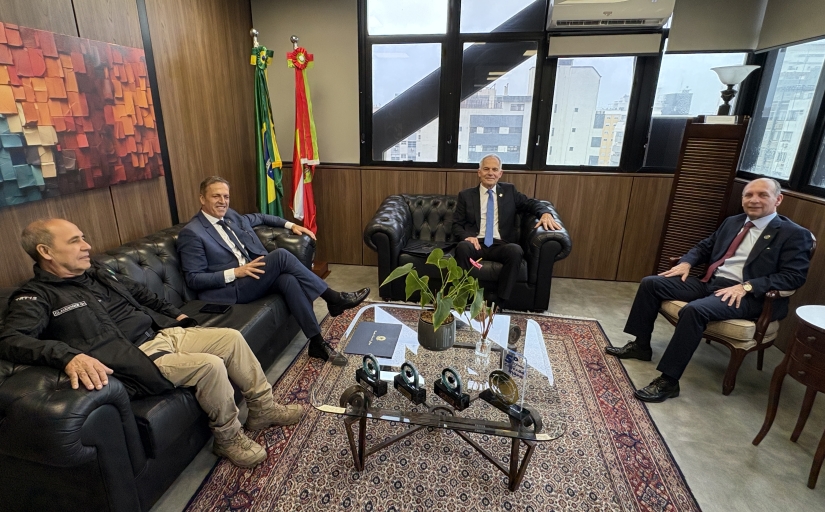 Três homens de terno e um com uniforme de segurança sentados em poltronas e cadeiras em uma sala arejada