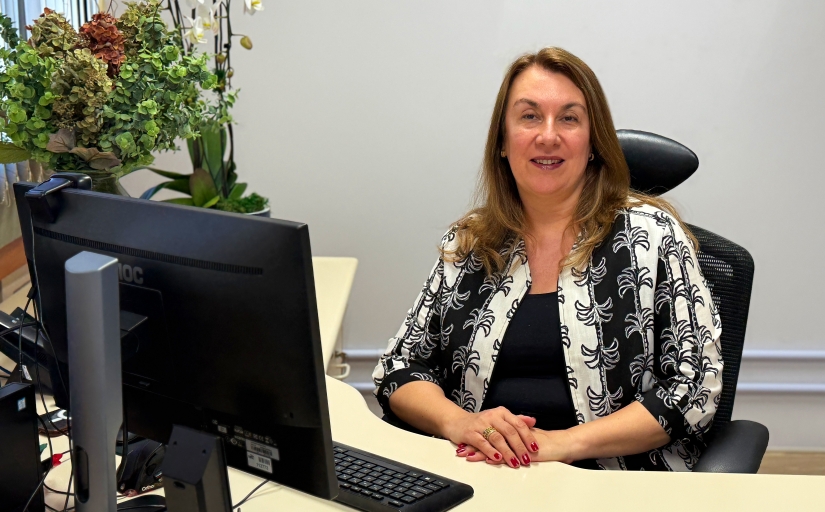 Mulher de pele branca e cabelos castanhos claros, sentada em um posto de trabalho, sorri para a câmera