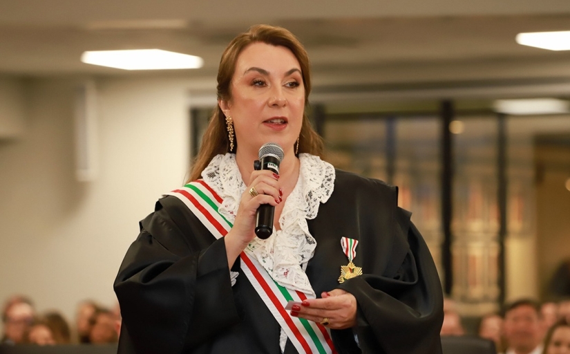 Mulher de toga e faixa fala ao microfone