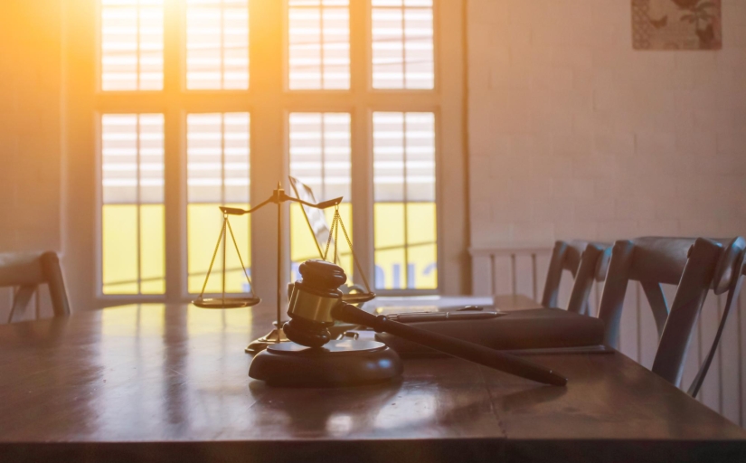 Foto de um martelo da justiça em cima de uma mesa de madeira. Ao fundo uma janela com o sol batendo como reflexo.