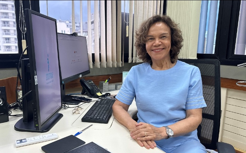 Mulher parda, madura, de conjunto azul claro e cabelos castanhos sentada à mesa de trabalho