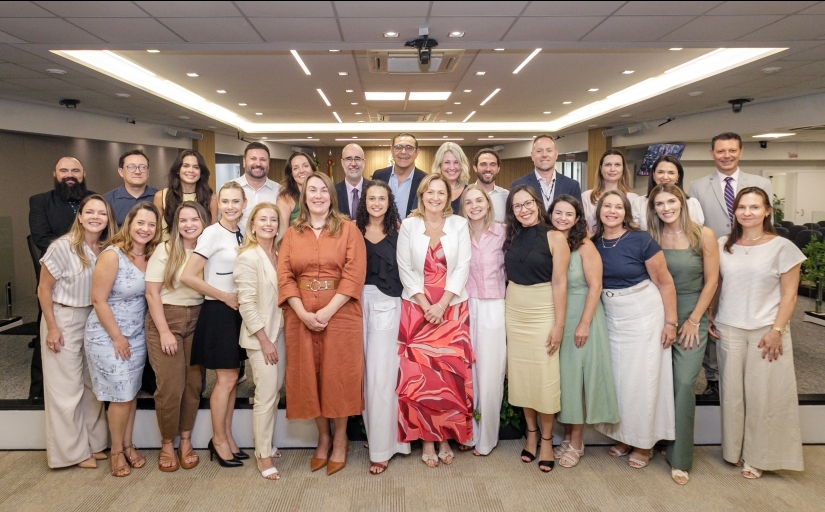 Grupo de 26 pessoas em pé, distribuídos em duas fileiras, posam para a foto em um auditório amplo