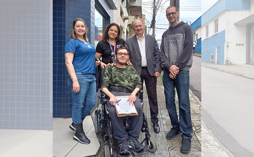 Cinco pessoas posam na calçada; jovem em cadeira de rodas segura prancheta.
