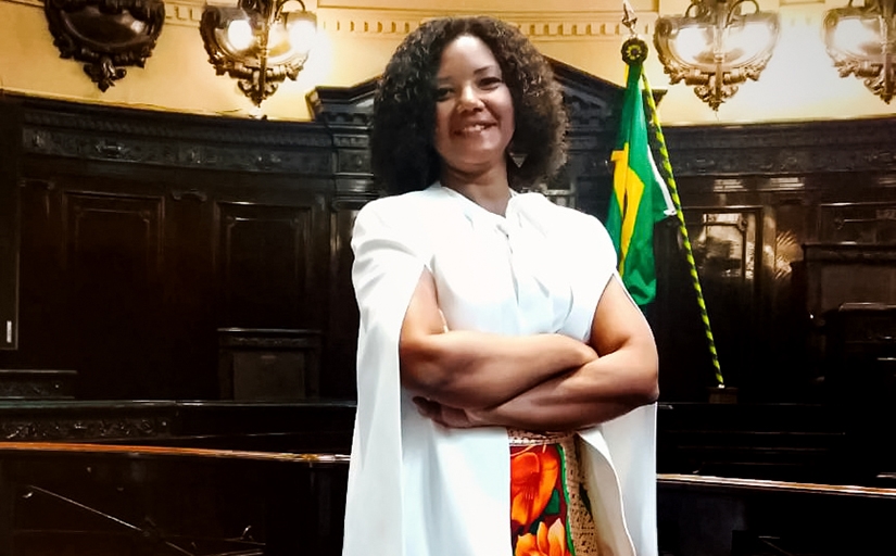 Mulher negra, de pé, de braços cruzados, em uma sala de julgamentos